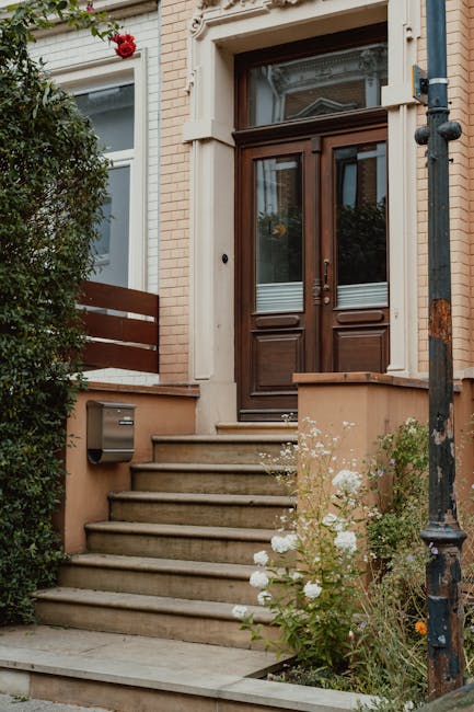 A quiet residential street with a narrow paved road flanked by well-maintained garden beds and tall, neatly trimmed hedges on the left side, and a variety of lush green trees and flowering shrubs on t